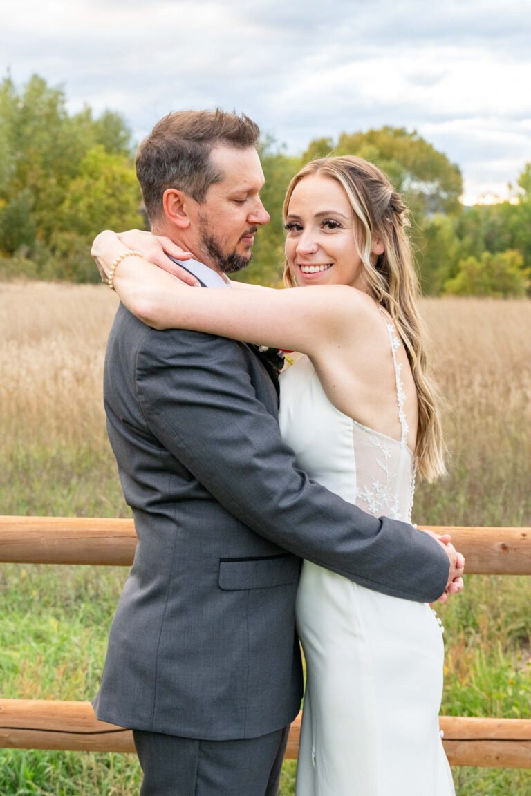 Leslie smiles while embracing Michael beside a wooden fence.