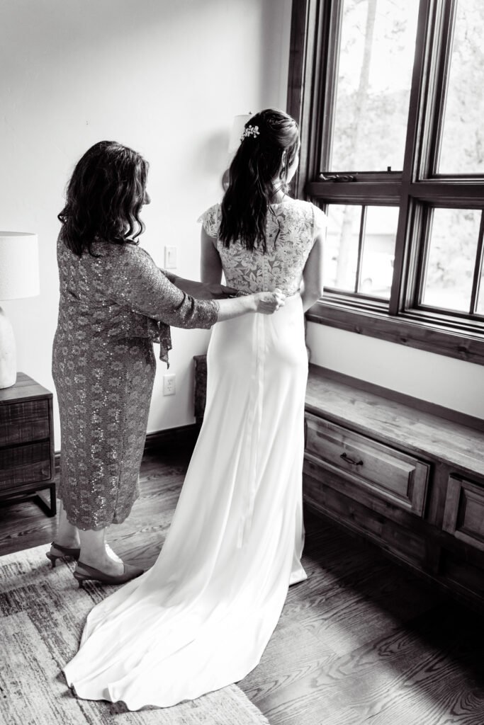 Black and white photograph of Nicole having her wedding dress fastened beside a window.