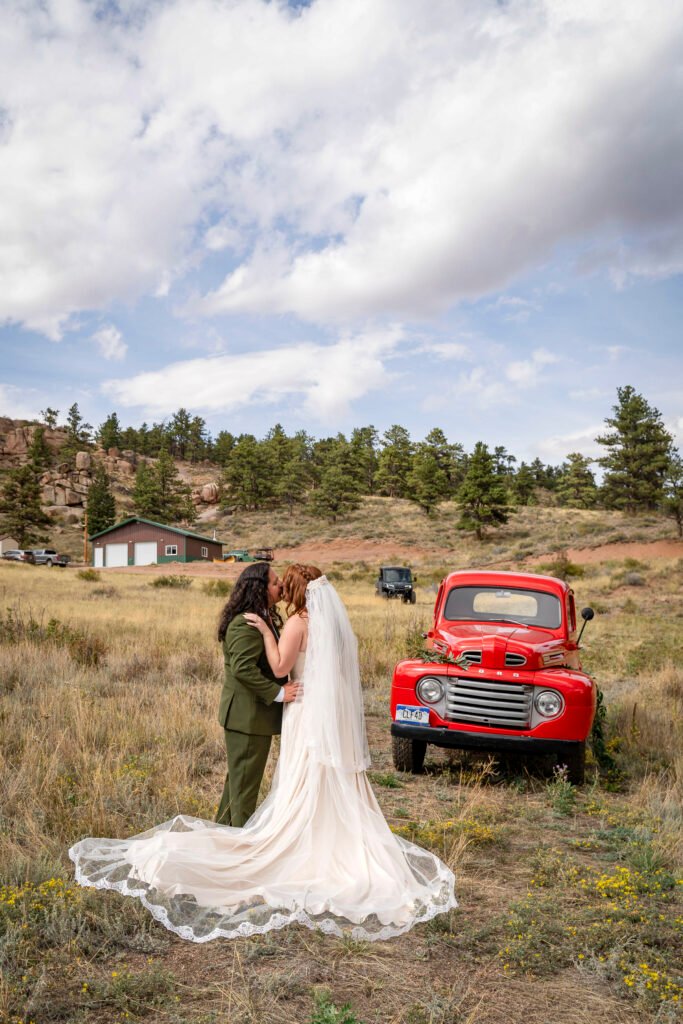Vince and Ash kiss beside a red vintage truck, with her wedding dress train spread across the meadow.