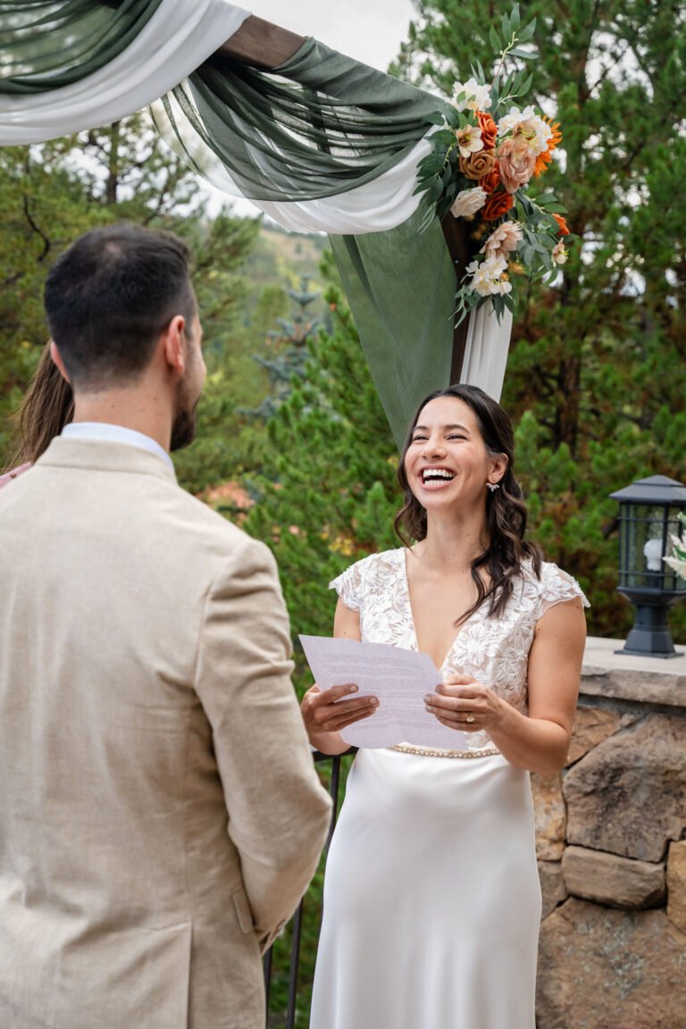 Nicole laughs while reading her wedding vows to Alex beneath a flower-decorated arch.