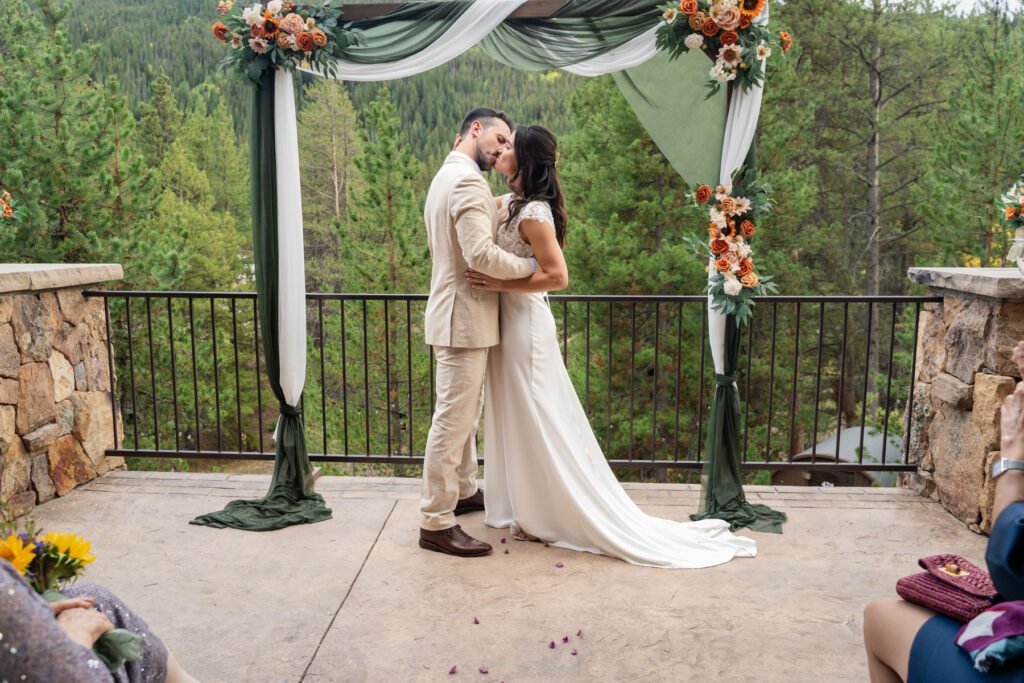 Alex and Nicole share their first kiss beneath a draped floral arch overlooking a forest.