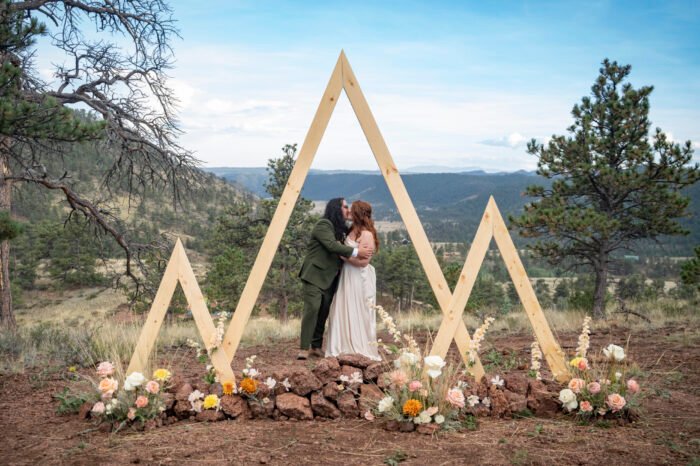 Vince and Ash embrace beneath a geometric wooden wedding arch decorated with flowers, with mountains behind them.