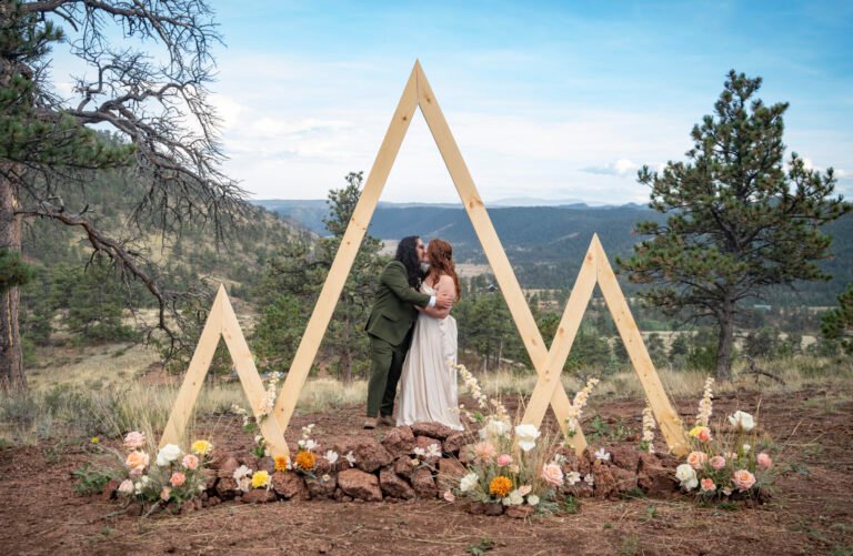 Vince and Ash embrace beneath a geometric wooden wedding arch decorated with flowers, with mountains behind them.