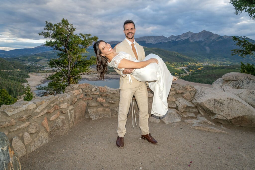 Alex carries a laughing Nicole in his arms with mountains and a lake behind them.
